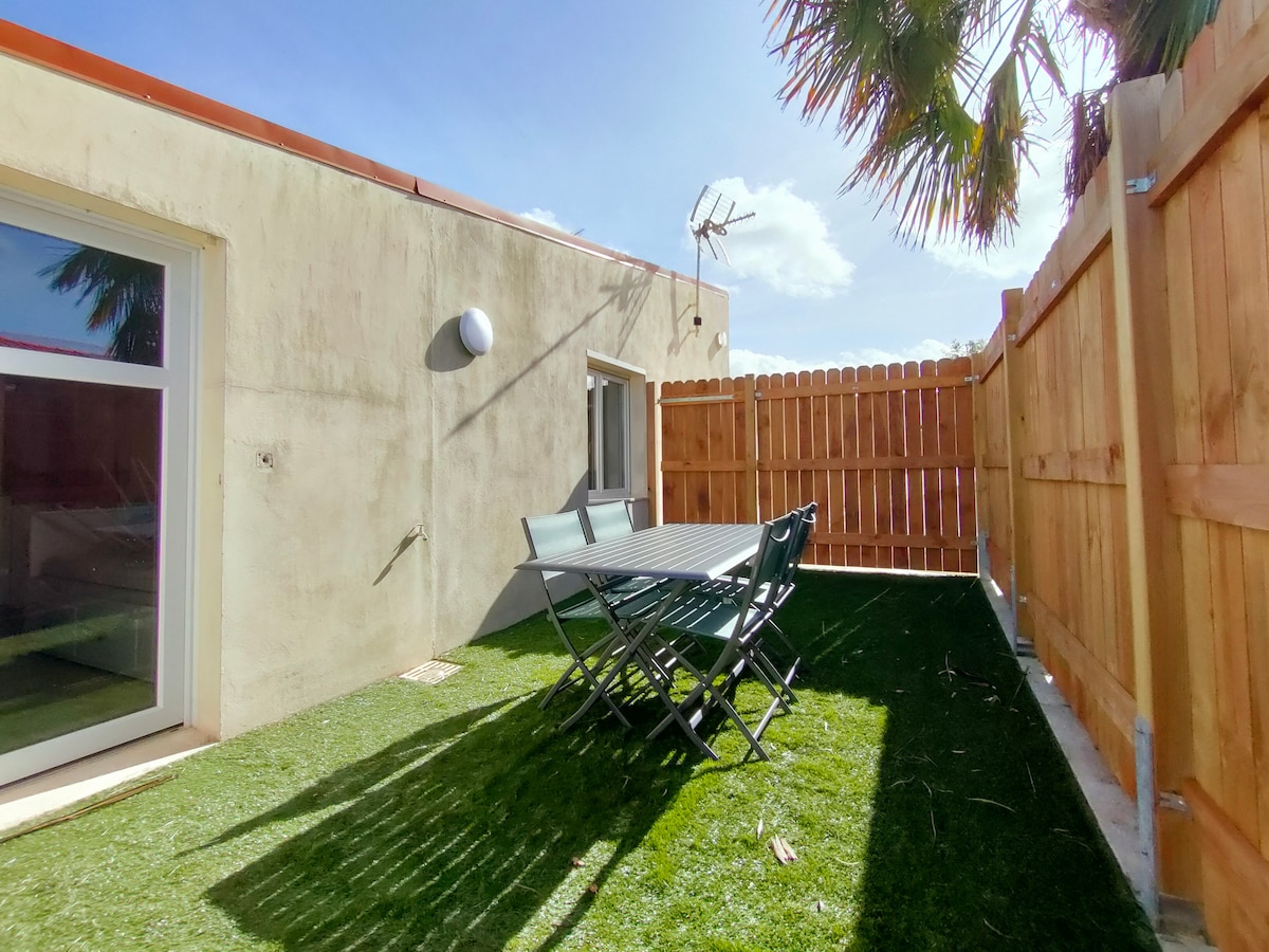 An outdoor terrace is presented with artificial grass, furnished with a rectangular table and six folding chairs. A wooden fence provides privacy, while a bright sky and palm leaves are visible in the background, creating a welcoming outdoor space.