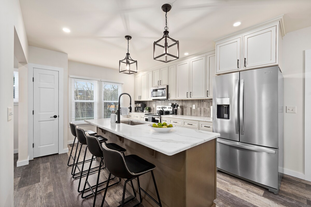 The kitchen features modern cabinetry and sleek stainless steel appliances. An island with bar stools provides seating and workspace. Large windows allow natural light to fill the space, highlighting the neutral tones and stylish fixtures throughout.