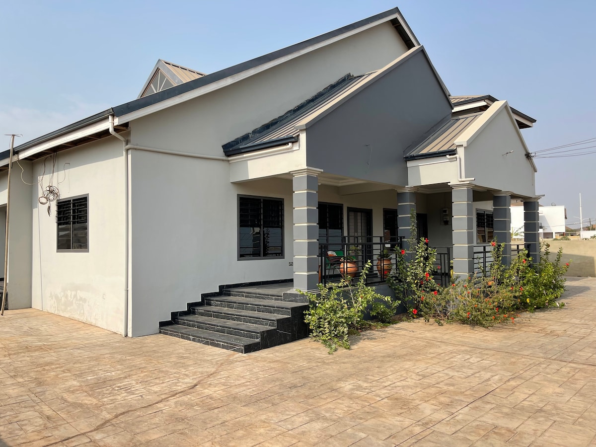 The exterior of the house features a modern design with a prominent entrance and steps leading to a covered porch. Surrounding greenery includes well-maintained plants, enhancing the property's appeal. Large windows allow natural light to enter, and the neutral color palette provides a welcoming look.