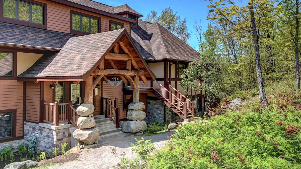 The exterior of the condo features a blend of natural stone and wood elements, accented by large decorative rocks near the entrance. Sturdy wooden stairs lead to the front door, surrounded by greenery and trees that enhance the peaceful setting.