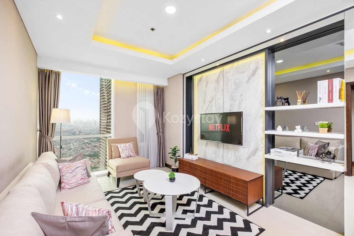 A modern living space features a soft beige sofa alongside a circular white coffee table. A sleek entertainment center displays a large TV with access to Netflix. Natural light floods in through floor-to-ceiling windows, offering expansive views of the city skyline.