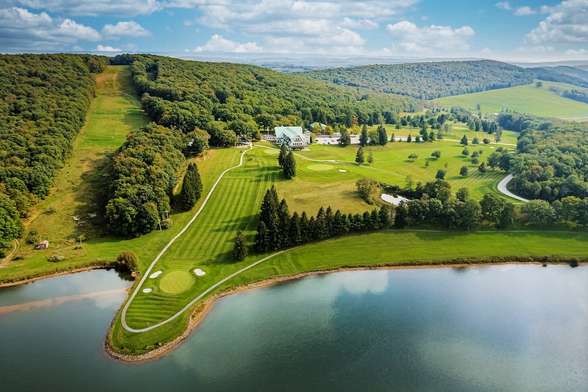 An aerial view showcases the expansive Alpine Lake Resort, surrounded by lush greenery and rolling hills. The manicured 18-hole golf course meanders along the lake's edge, reflecting the landscape. The scene captures the serene connection between nature and the resort, inviting guests for outdoor activities.