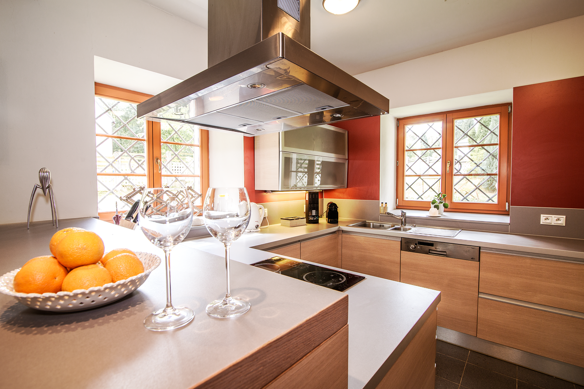 A well-equipped kitchen features wooden cabinetry and modern appliances. A central island is topped with a bowl of oranges, while two crystal glasses stand ready for use. Natural light filters through diamond-pane windows, creating a warm atmosphere.