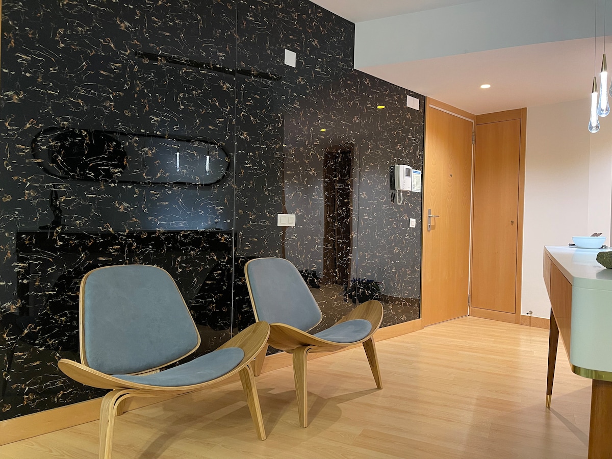 Two contemporary chairs with light blue upholstery and wooden frames are positioned in a welcoming corner. A polished black marble wall with gold veining serves as a backdrop, while soft lighting from overhead fixtures enhances the modern design.