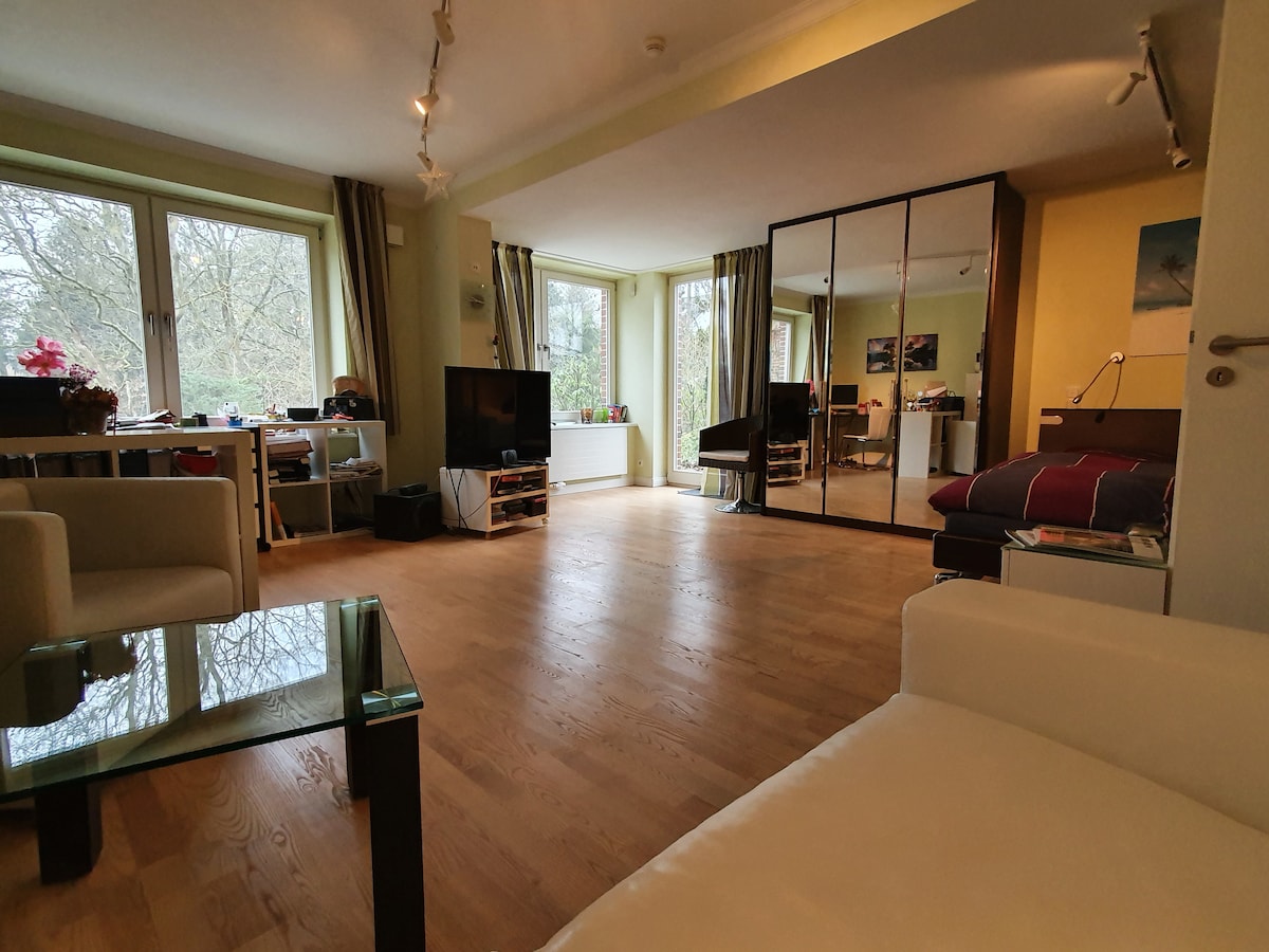 A spacious living area features large windows that provide natural light and views of the garden. A comfortable seating arrangement is visible, along with a flat-screen television. A dining space can be seen in the background, complemented by modern furnishings and wooden flooring.