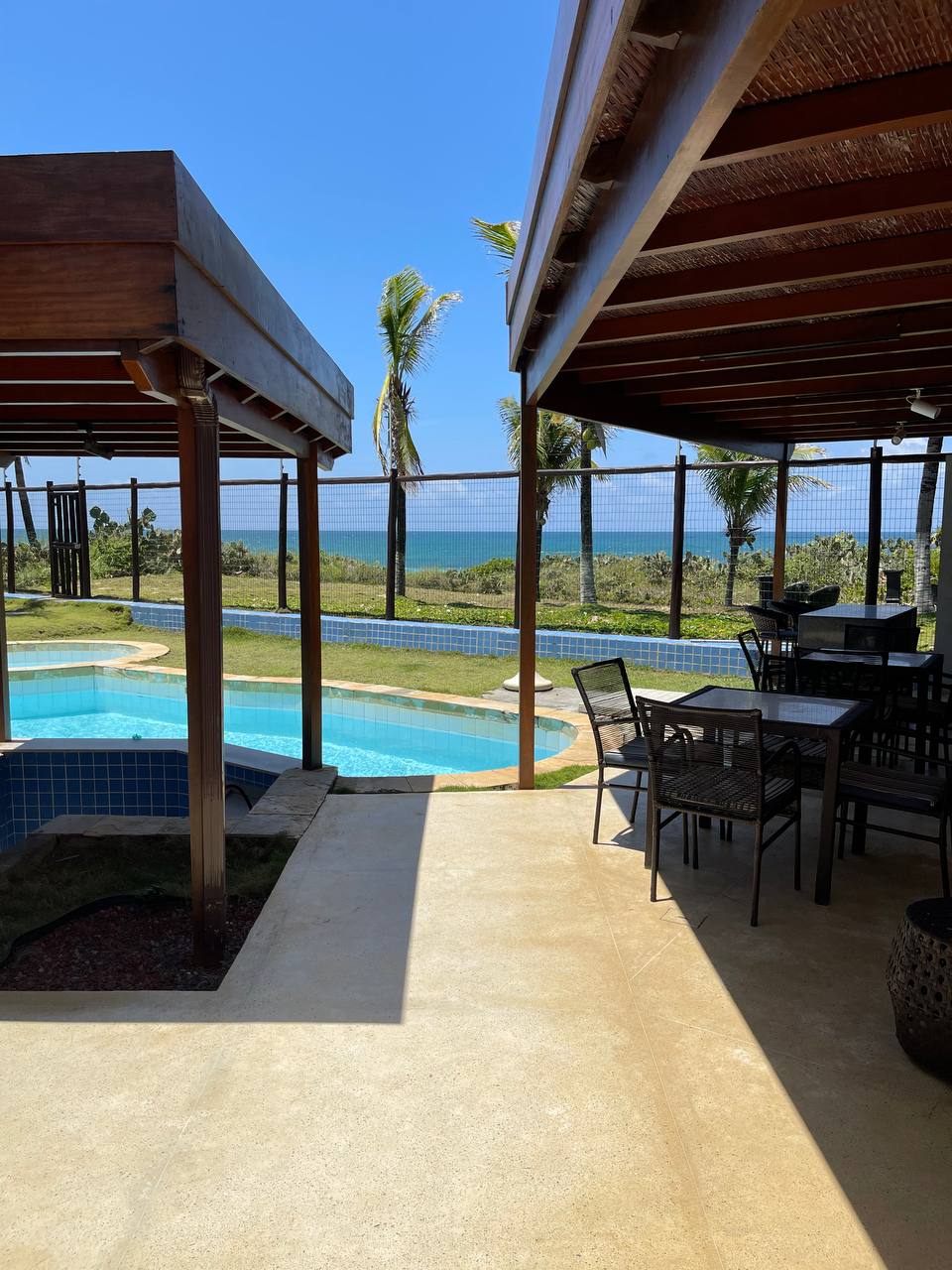 A covered outdoor area featuring a dining table and chairs overlooks a spacious pool and scenic views of the landscape and sea. The sun reflects on the water, and palm trees sway gently in the background, contributing to the serene environment.