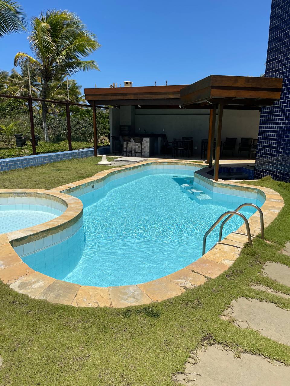 The image features a refreshing pool, surrounded by neatly trimmed grass and natural stone pathways. A shaded sitting area is visible to the rear, along with tall palm trees in the background. Sunlight reflects on the clear water, creating a serene atmosphere.