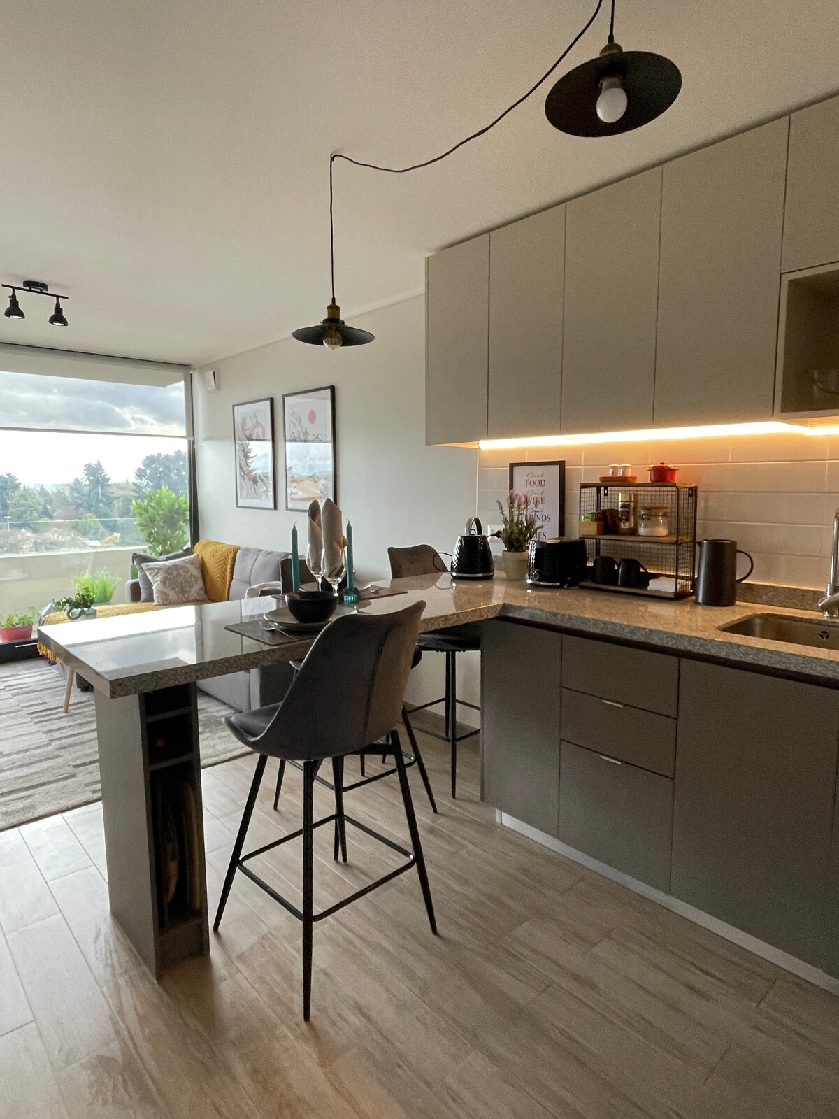 A modern kitchen area features sleek cabinetry and a granite countertop with bar seating. Natural light fills the space through large windows, illuminating a cozy seating area with armchairs. Decorative plants add greenery, enhancing the overall atmosphere of the room.