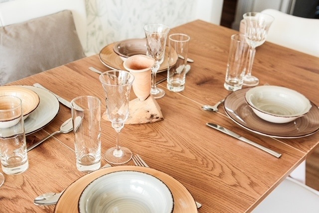 A wooden dining table is set for a meal, featuring an arrangement of various plates and glassware. Two types of plates provide a contrasting appearance, with transparent and patterned glasses placed alongside cutlery. A small decorative item is centered on the table, contributing to the overall presentation.
