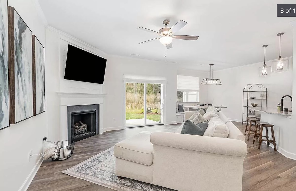 A bright living area showcases a spacious sectional sofa with plush cushions facing a flat-screen television mounted on a white wall. A fireplace is positioned below the TV. Large sliding doors open to a private patio, allowing natural light to fill the space.