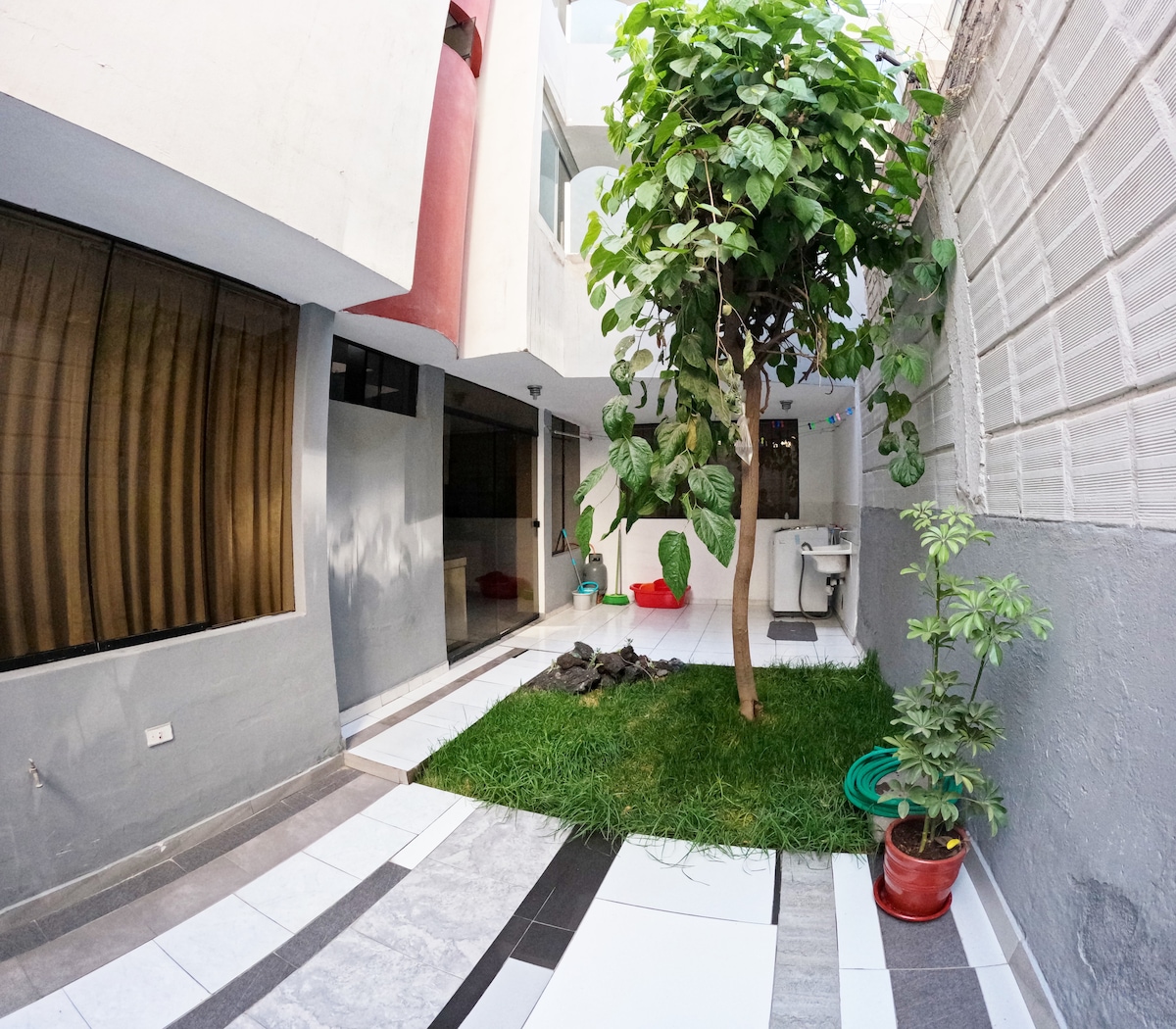 A tranquil outdoor patio area features a well-maintained garden with a central tree surrounded by green grass. Sunlight filters through the area, highlighting the patio's tiled surfaces and providing a relaxing space with planters and a laundry area in the background.