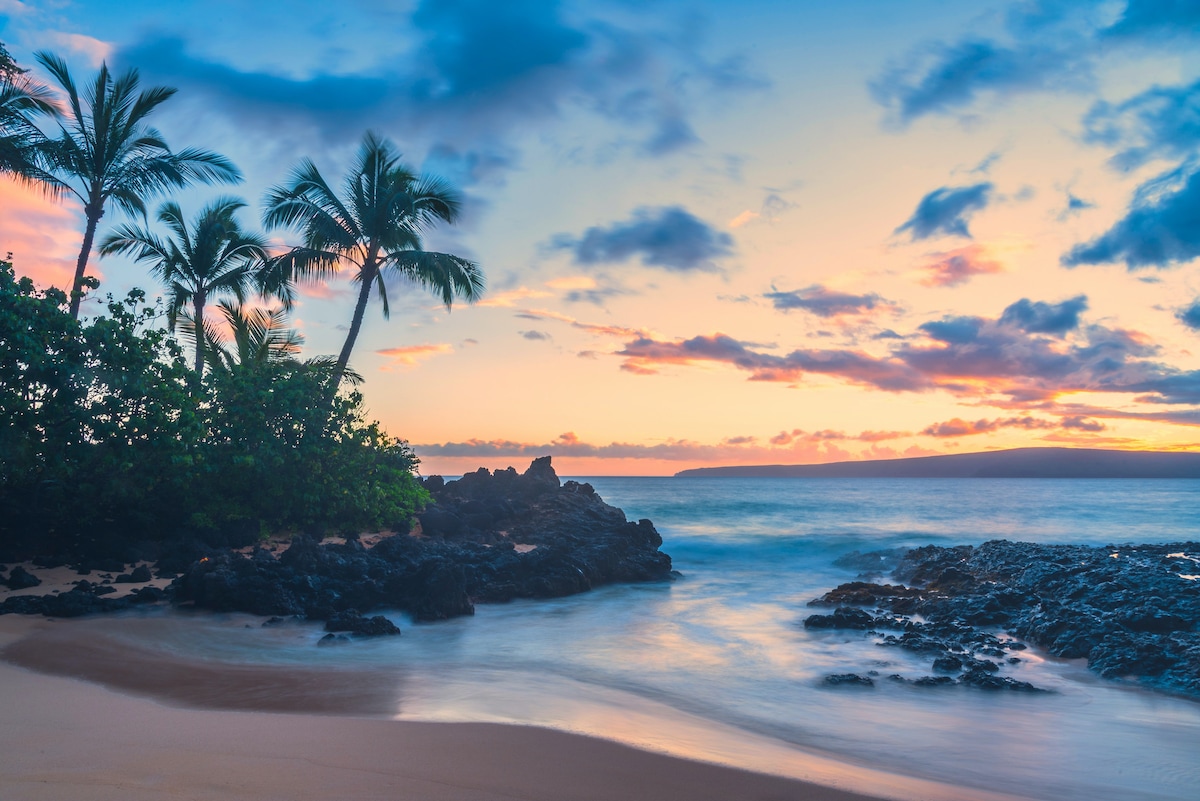 The image captures a serene beach scene at sunset, featuring gentle waves lapping against rocky outcrops. Lush green palm trees frame the view, while soft colors of orange, pink, and blue fill the sky. The distant outline of an island is visible on the horizon.
