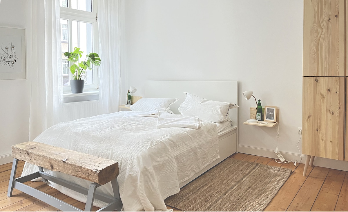 A spacious bedroom features a comfortable bed dressed in white linens. Natural light streams in through a large window, illuminating simple decor. A green plant on the windowsill adds a touch of nature. Wooden elements, including a bench and wardrobe, complement the light wooden flooring.