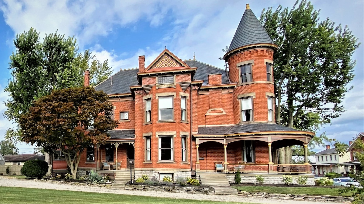 The exterior of a grand Queen Anne-style house is highlighted, showcasing its intricate brickwork and distinctive turret. A spacious porch wraps around the front, bordered by carefully tended landscaping, including a vibrant tree and well-manicured flower beds, under a partly cloudy sky.