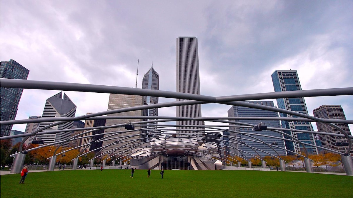 A modern outdoor park features a green lawn under a dramatic sky. The skyline of tall city buildings creates a backdrop, with a distinctive architectural structure at the center. The space is open and accessible, inviting visitors to enjoy the surrounding urban landscape.