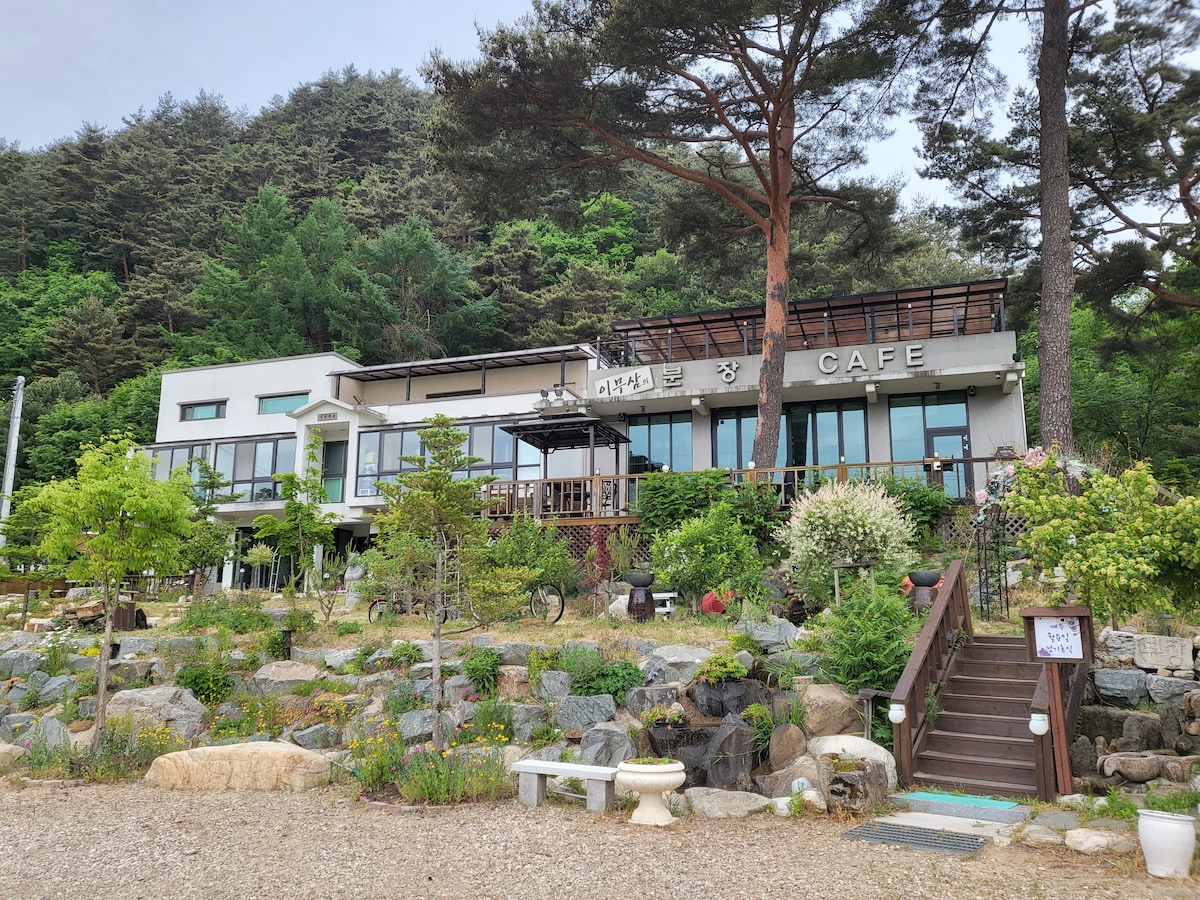 The exterior of a modern cafe is shown, surrounded by a landscaped garden featuring various shrubs and trees. Large windows allow natural light to fill the interior, while a spacious deck offers outdoor seating. Stone pathways guide visitors through the green area.