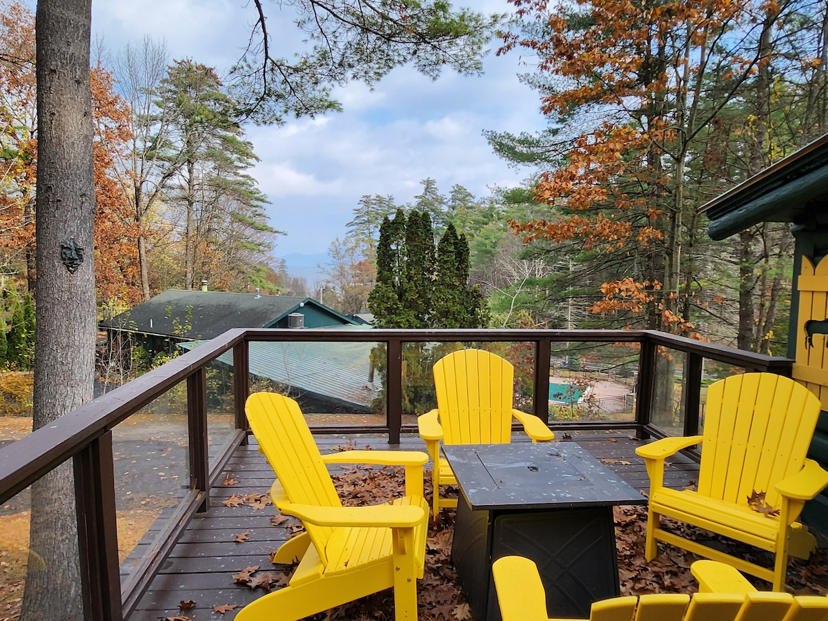 A spacious deck is shown with four bright yellow adirondack chairs surrounding a central black table. Leaves in autumn hues cover the wooden floor, while trees with vibrant foliage surround the area, creating a serene outdoor space. Distant mountains are faintly visible in the background.
