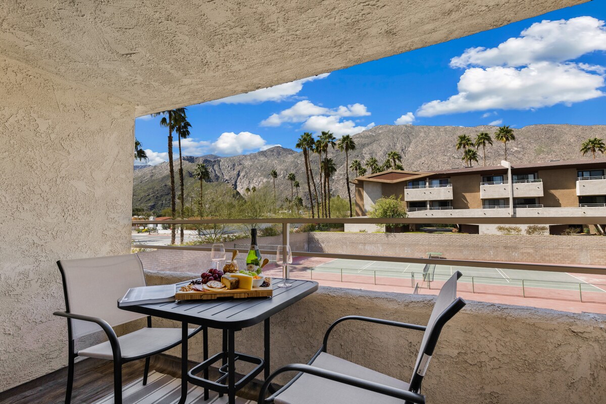 A private balcony offers a dining table with two chairs, set for outdoor enjoyment. The view showcases lush palm trees and distant mountains under a blue sky, while tennis courts are visible in the background.