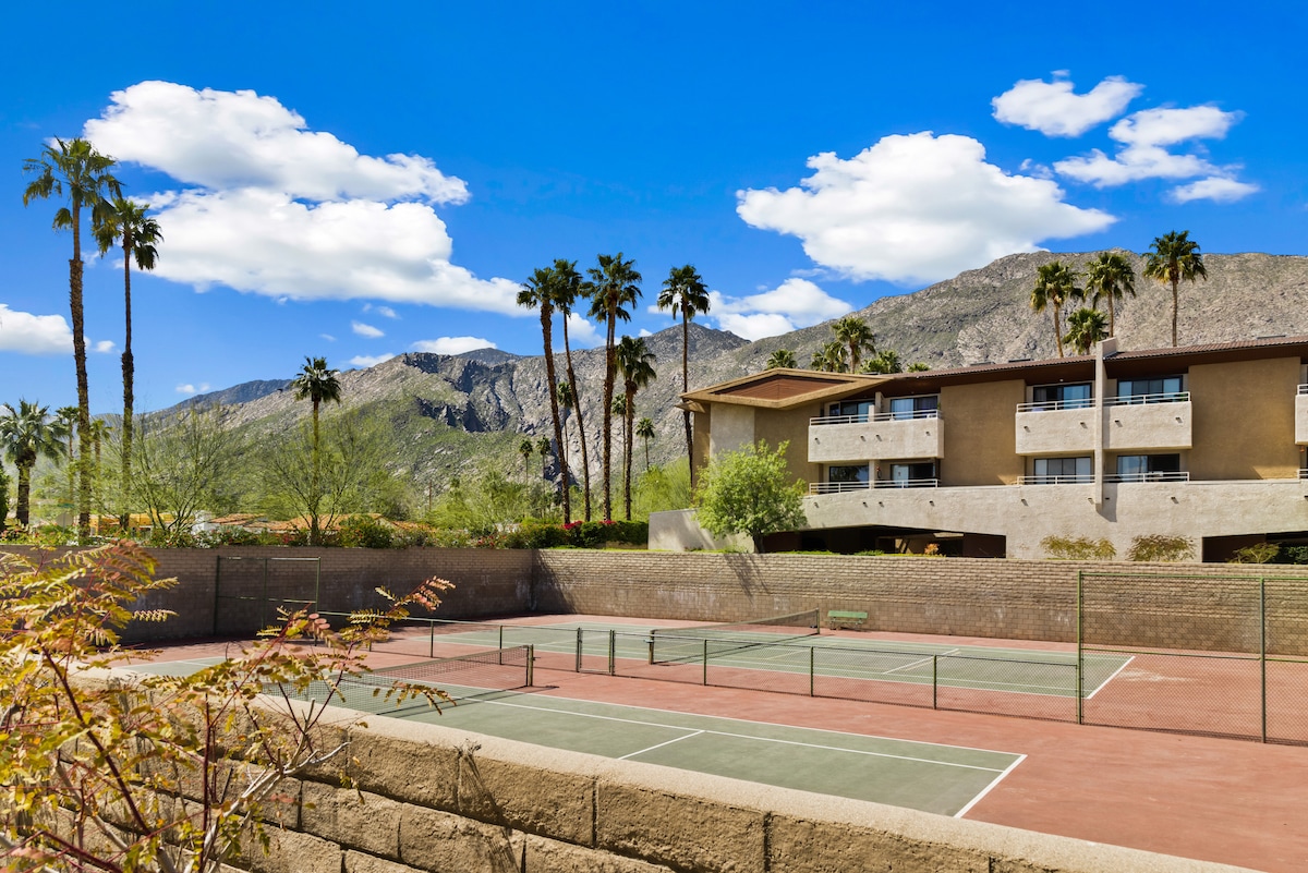 Two well-maintained tennis courts are framed by lush palm trees and mountain views in the background. The clear blue sky enhances the vibrant outdoor setting, showcasing the condo community's active atmosphere. The courts feature simple markings and are ready for play, inviting guests to enjoy outdoor activities.