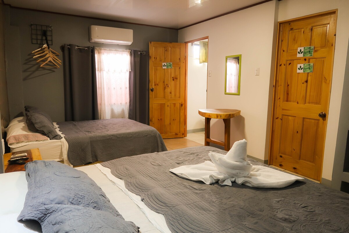 The room contains two beds with gray bedspreads, framed by wooden accents. Light enters through a curtained window. A small round table and wooden doors add to the simplicity and functionality of the space, complemented by air conditioning visible on the wall.