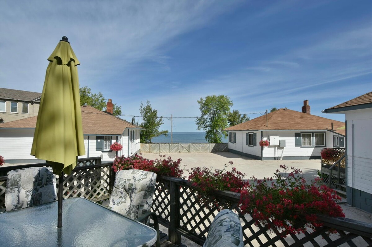 An outdoor deck features a glass table surrounded by floral-patterned chairs. A green umbrella provides shade, while vibrant hanging flowers add color to the space. A serene view of blue skies and the lake is visible in the background, along with neighboring cottages.