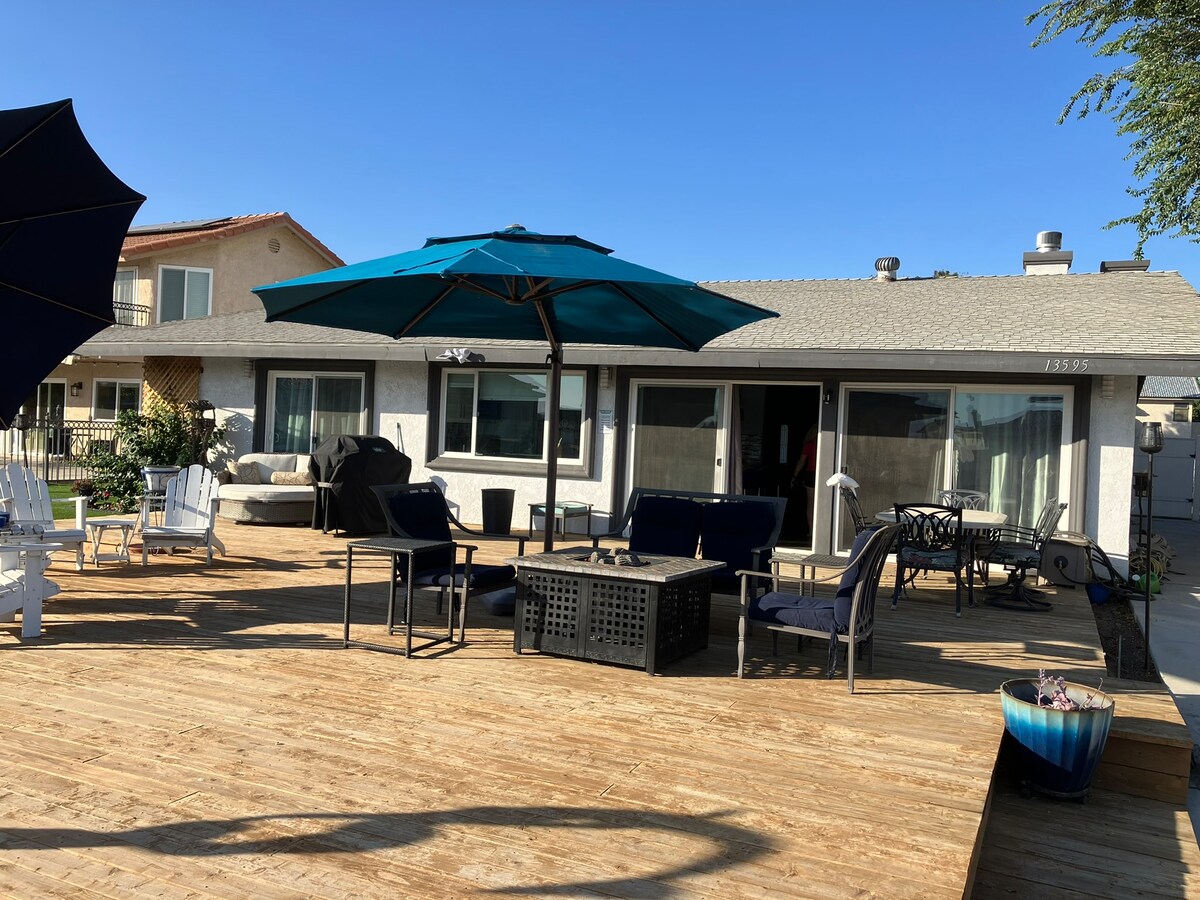A spacious wooden deck features multiple seating areas, including lounge chairs and a dining table. Large umbrellas provide shade. The home's glass doors open to the interior, enhancing the connection between indoor and outdoor spaces.