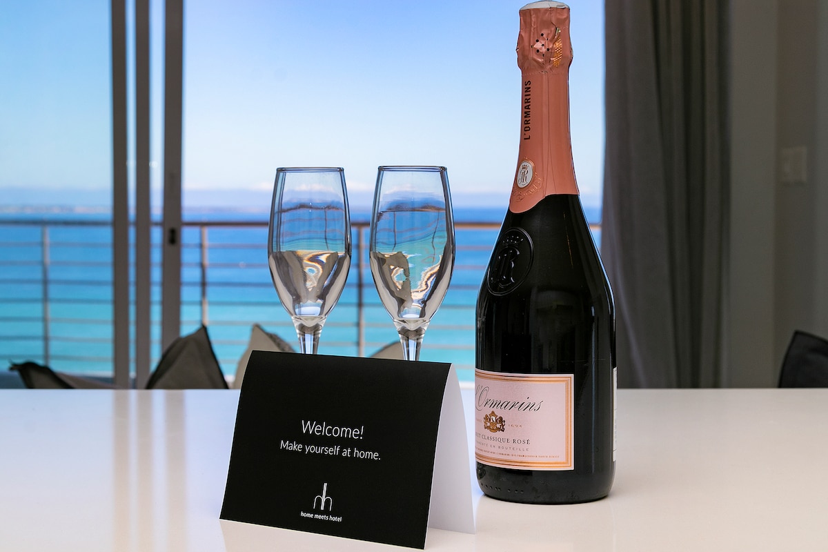 A bottle of sparkling rosé wine is positioned next to two elegant glasses on a polished countertop. A welcome card invites guests to feel at home, with a stunning ocean view visible through the glass railing in the background.