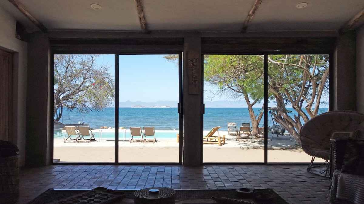 A panoramic view of the lake is framed by sliding glass doors, showcasing a tranquil poolside area with sun loungers. The blue waters of the lake reflect the clear sky, while lush greenery and trees provide shade in the foreground.