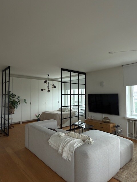 A modern living area features a light-colored sofa with a cozy throw draped over one end. Behind a stylish partition, a bedroom is visible, complemented by large windows. A flat-screen TV is mounted on a wall, and minimalist wood furniture enhances the space.