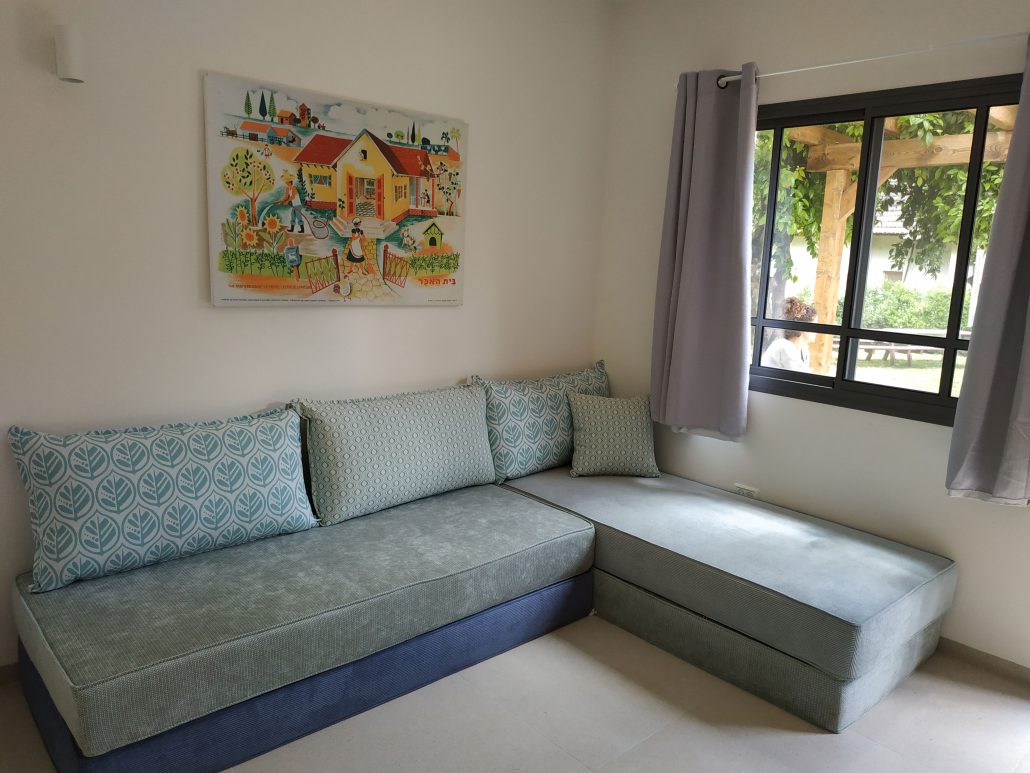 A comfortable seating area features a spacious sectional couch adorned with patterned cushions. Natural light enters through a large window, framed with light curtains, illuminating a colorful wall art piece displayed above the couch.