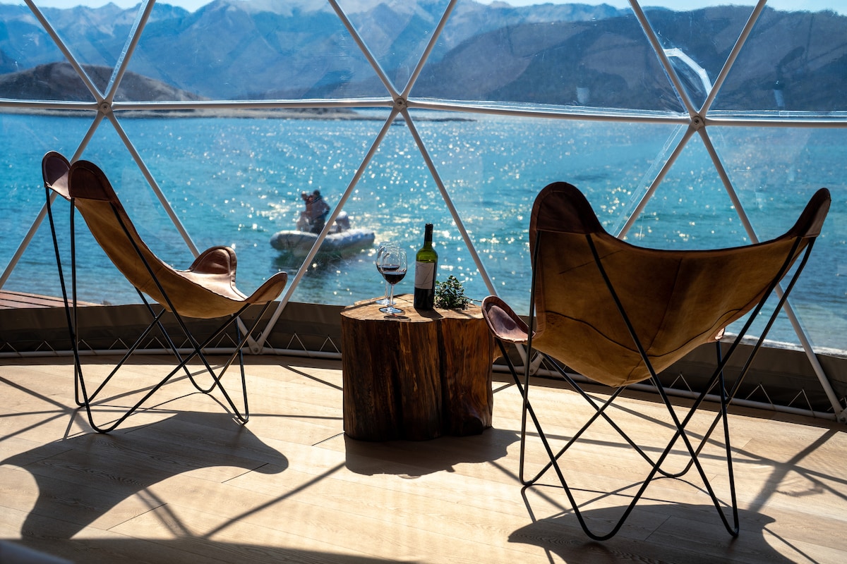Two butterfly chairs are positioned near a wooden side table and overlook the shimmering water of Lake Nahuel Huapi. A bottle of wine and a glass sit on the table, framed by a panoramic view of the mountains in the distance.