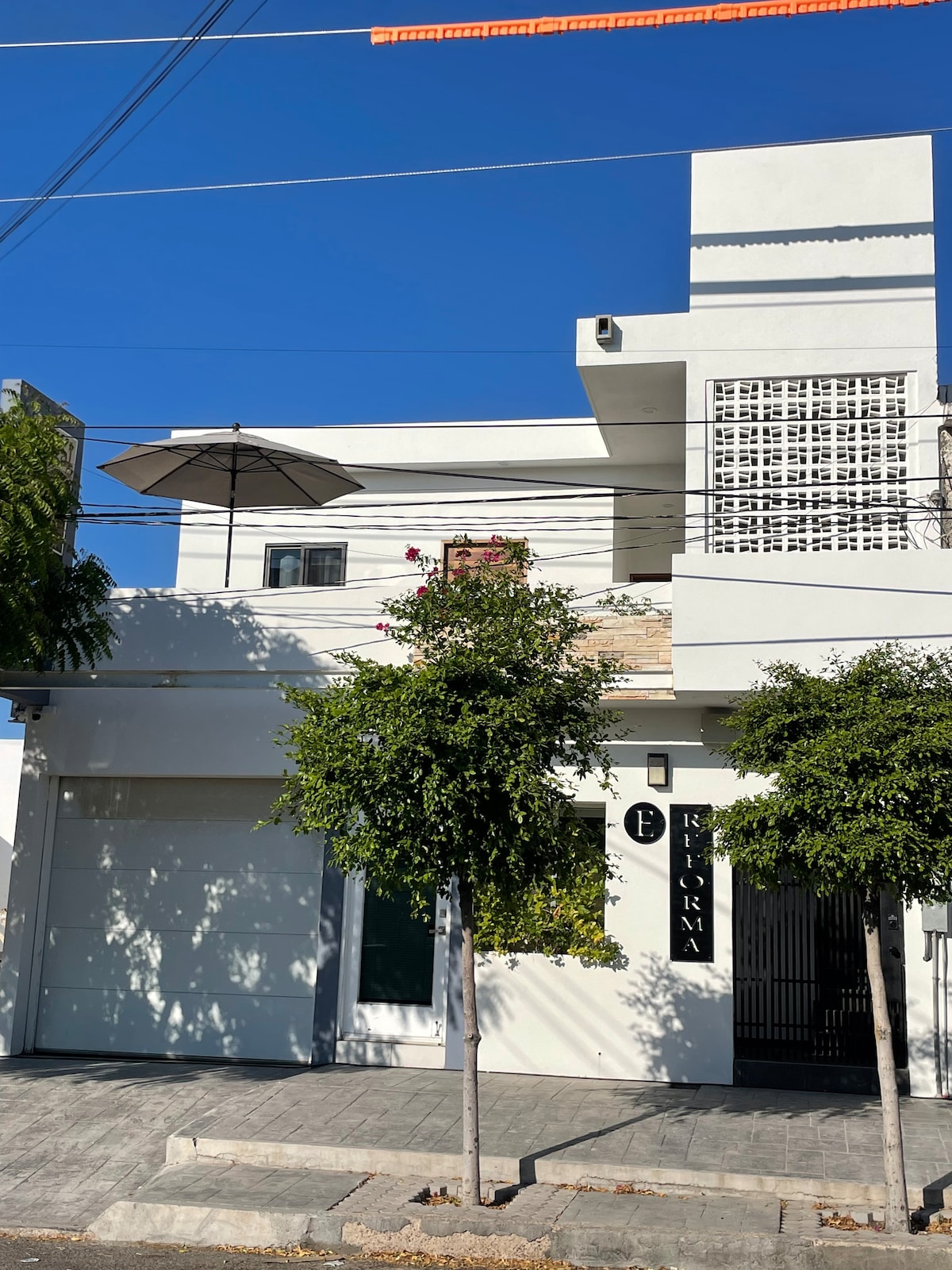 The exterior of a modern building features a clean white facade, complemented by a vibrant greenery. A large umbrella is seen atop the terrace, providing shade. Decorative tile work can be noted along with clear blue skies in the background.