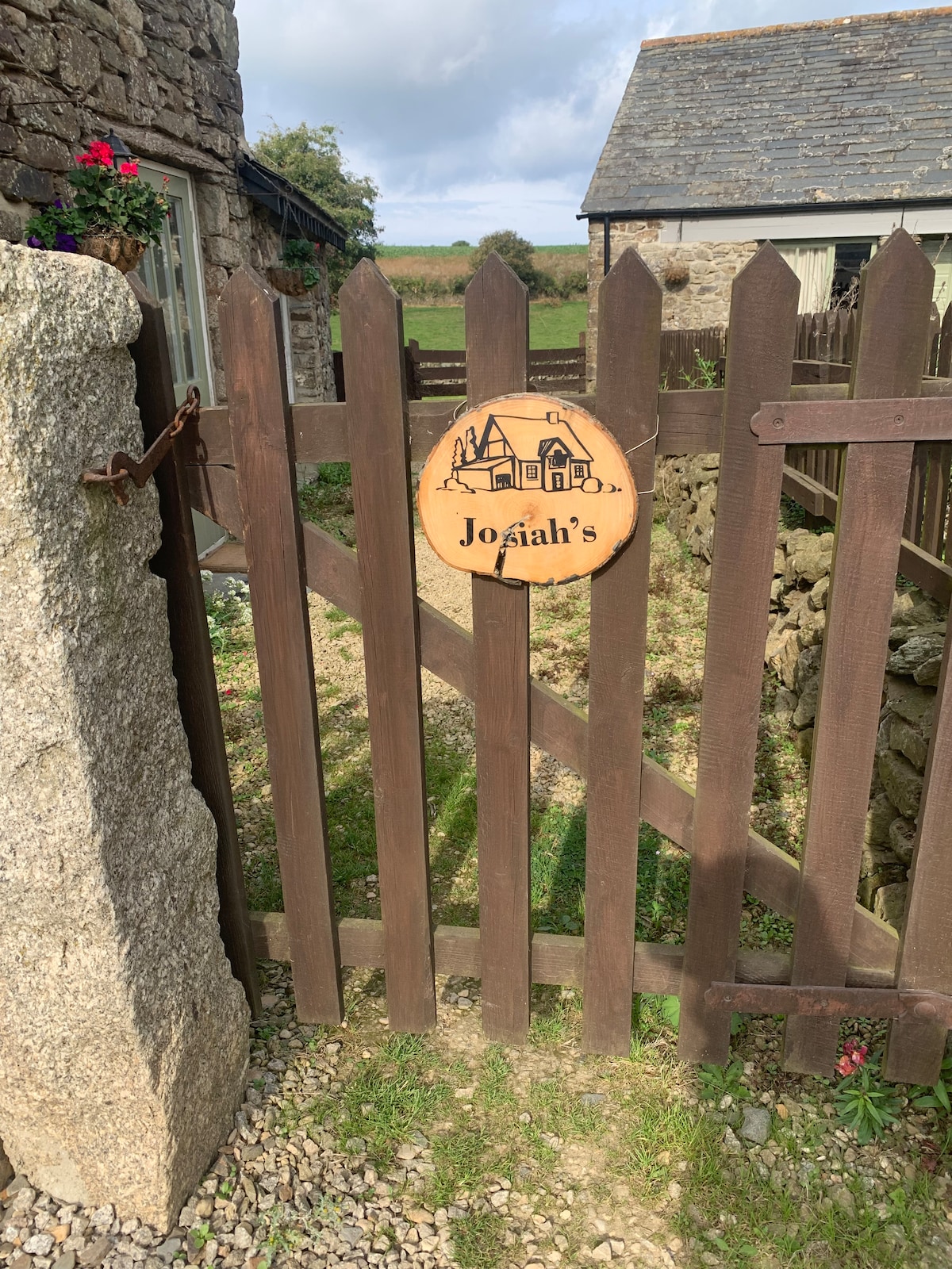 A wooden gate, painted in dark brown, is depicted with a sign displaying the name 'Josiah's.' The entrance is surrounded by a stone wall and a grassy area, leading to an inviting view of the landscape beyond.