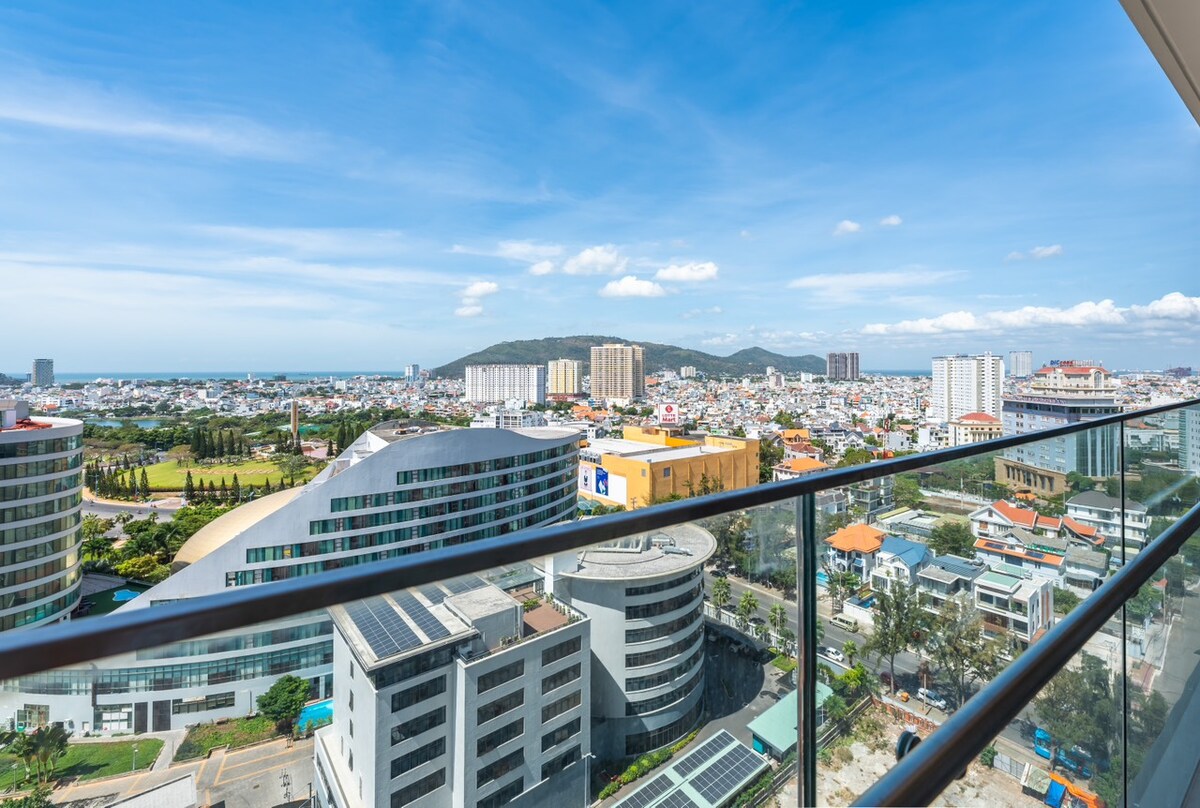 A panoramic view from the balcony captures the vibrant cityscape, mountains, and coastline. The clear sky enhances visibility, showcasing various buildings and green areas below. The glass railing offers an unobstructed sightline, emphasizing the spaciousness of the outdoor area.
