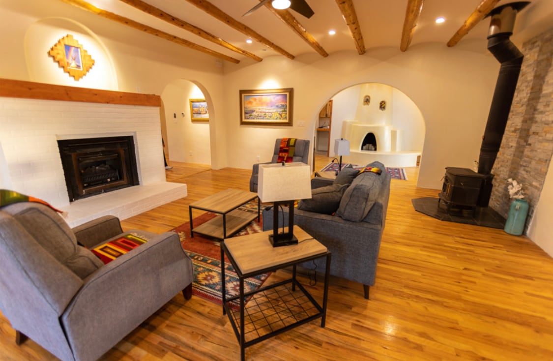 A spacious living area features light wood flooring and traditional viga beamed ceilings. Comfortable seating is arranged around a central coffee table, with a fireplace and a wood-stove seated against a textured stone wall. Art pieces hang on the walls, adding a touch of local charm.