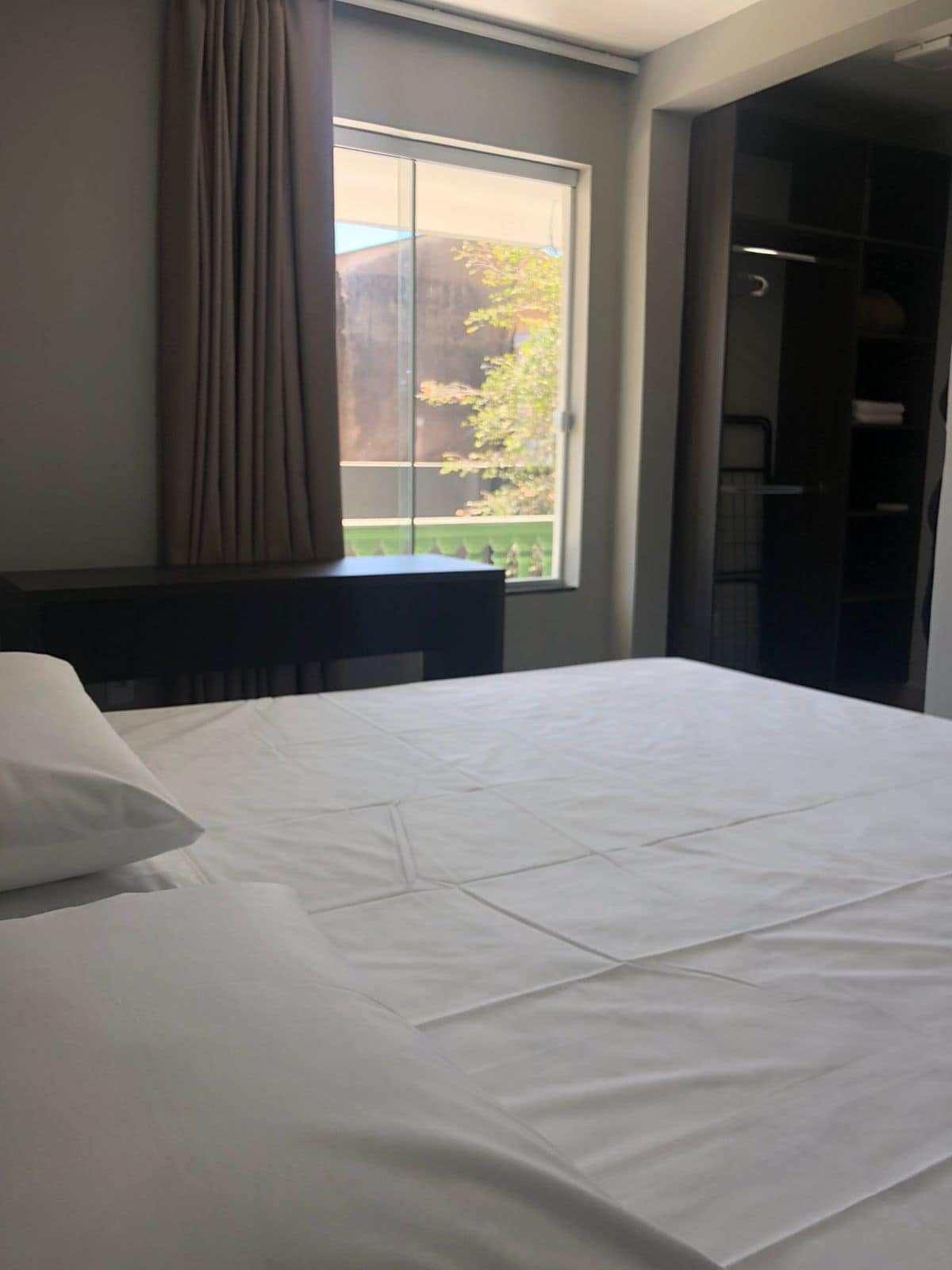 A cozy sleeping area features a spacious bed with crisp white linens. Natural light enters through a large window dressed with curtains, illuminating the room. A dark wood desk is positioned beside the bed, and a closet space can be seen in the background.