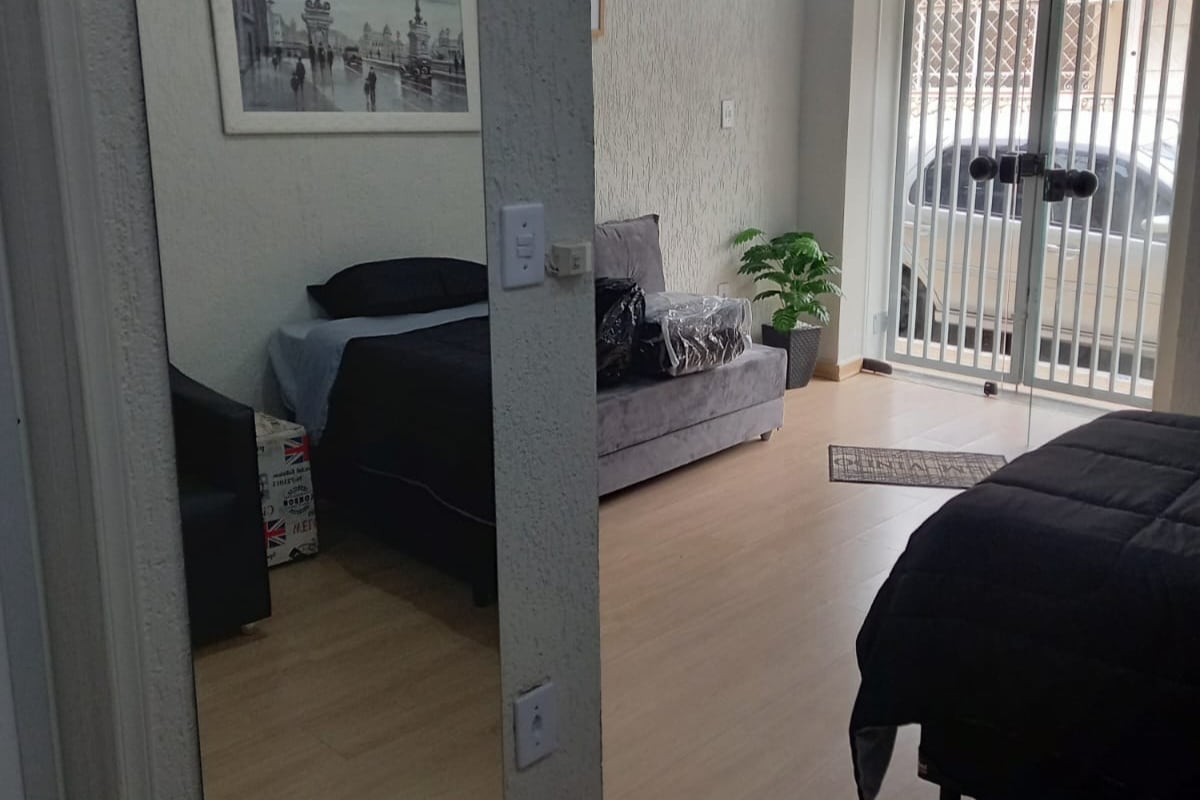 The image displays a cozy interior view of the flat, featuring a spacious bed with dark bedding and a grey sofa. A small plant adds a touch of greenery, while a doorway leads to the light-filled common space with sliding glass doors.