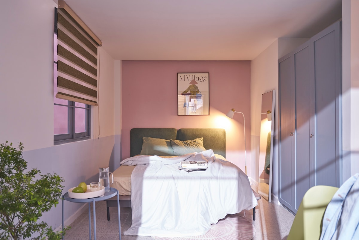 A comfortable bedroom is illuminated by warm lighting, featuring a spacious bed with light bedding and decorative pillows. A small side table holds a glass of water and an apple. A piece of artwork is hung above the bed, and a full-length mirror reflects the room's pastel tones.