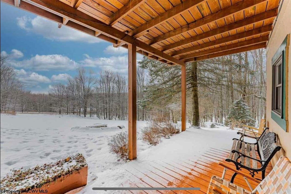 A spacious covered porch features wooden beams and a wooden floor, extending towards a snow-covered landscape. Two benches provide seating in front of a scenic view of trees and open space, creating a peaceful outdoor retreat.