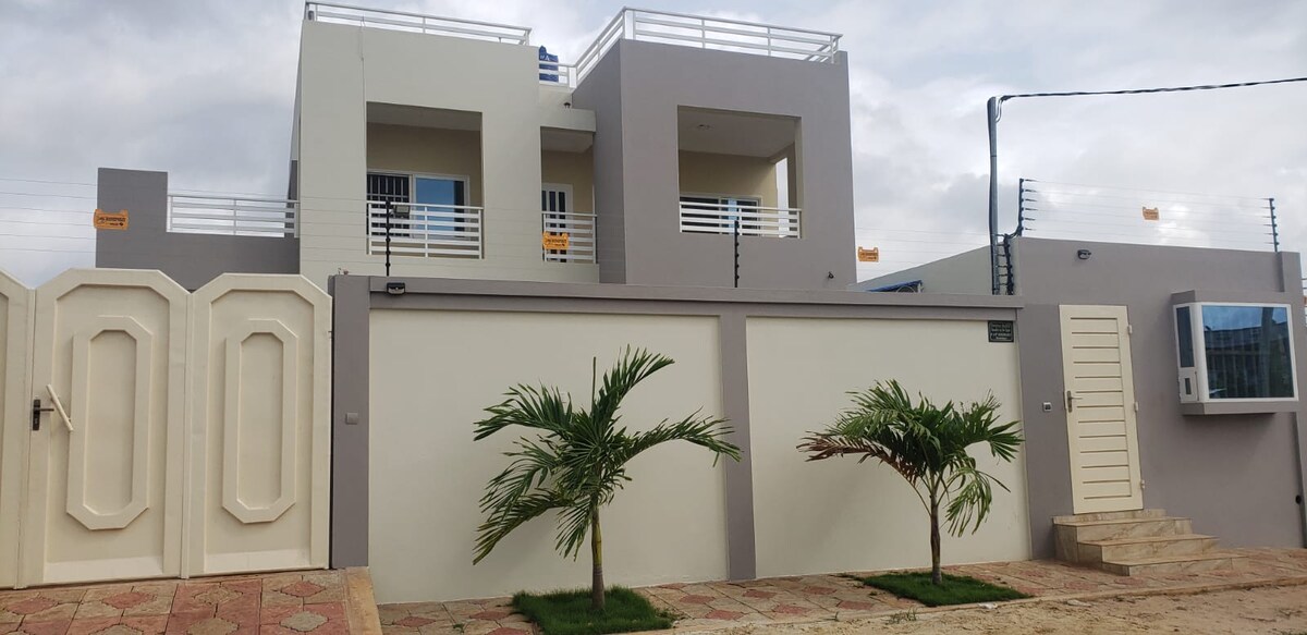 The villa showcases a modern exterior with a two-story design. Light-colored walls are complemented by a stylish entrance gate. Two palm trees are planted in front, enhancing the welcoming appearance. A balcony is visible on the upper level, providing additional outdoor space.