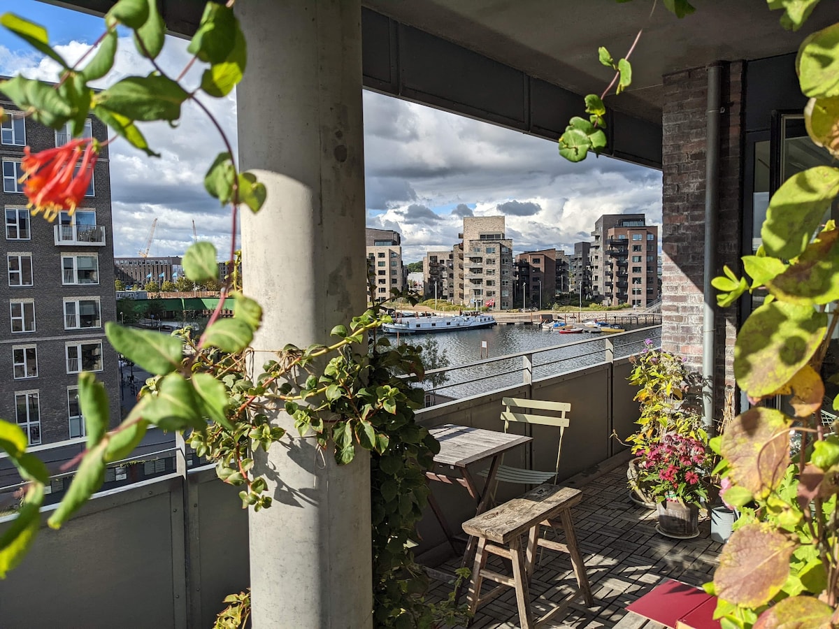 A balcony is framed by lush greenery, featuring a small wooden table and a folding chair. Below, a scenic view of charming canal-side buildings reflects life by the water. Sunny skies and scattered clouds provide a serene backdrop to the vibrant foliage.