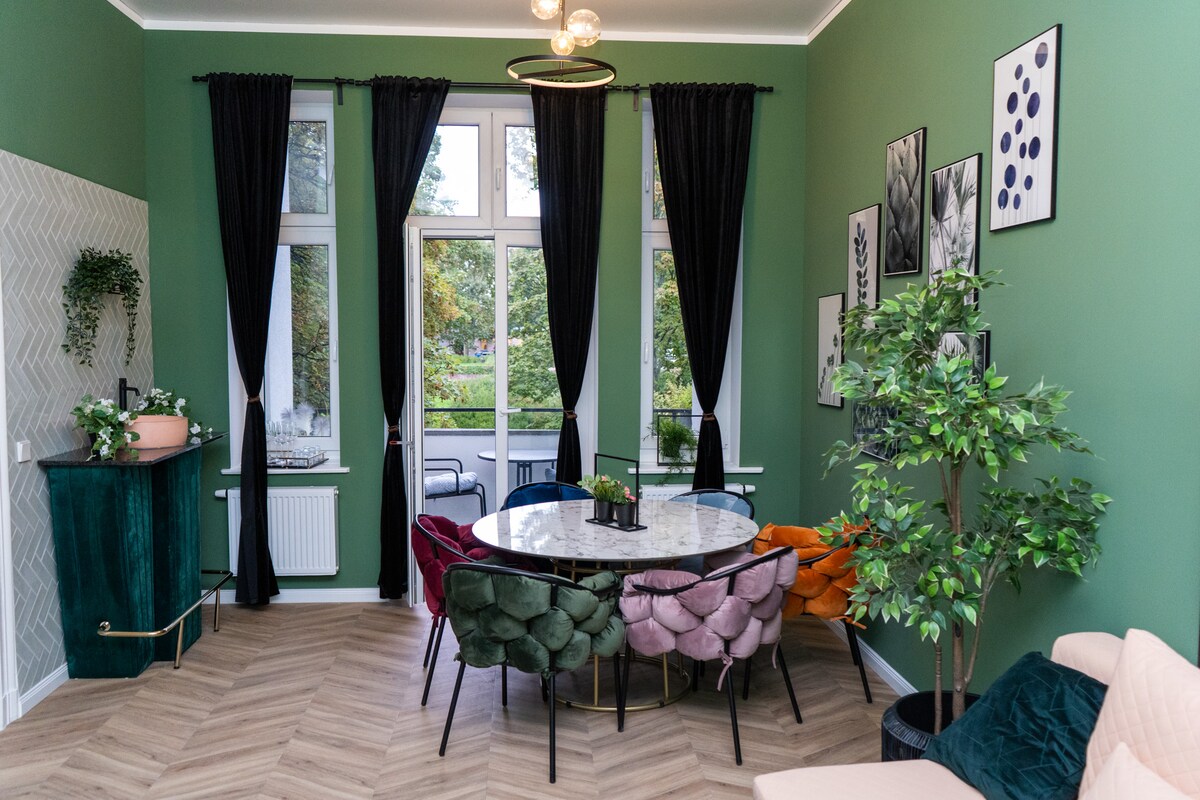 The living room features large windows framed by black curtains, allowing natural light to brighten the space. A round marble dining table with colorful, upholstered chairs contrasts the green walls. Indoor plants and wall art add vibrancy to the inviting atmosphere.
