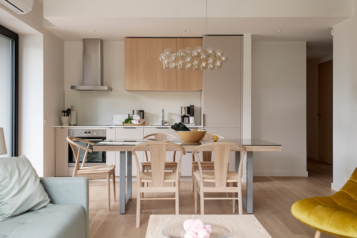 A modern kitchen and dining area features an elegant table surrounded by four wooden chairs. A simple bowl is centered on the table, with light-colored cabinetry and appliances visible in the background. Natural light streams through large windows, illuminating the open layout.