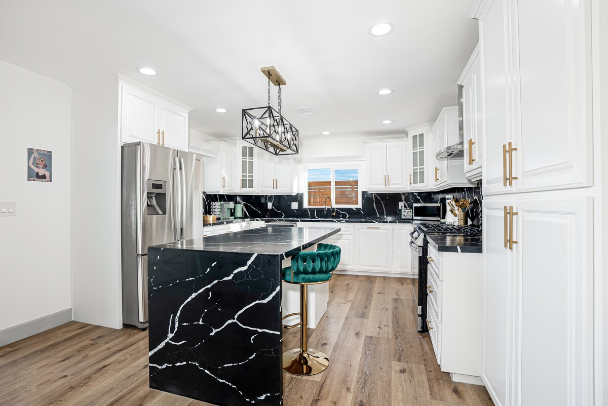 A spacious kitchen features striking black marble countertops with white veining, complemented by modern white cabinetry. Stainless steel appliances are visible, including a refrigerator and stove. An island with seating is highlighted by a pendant light, and natural light filters in through a nearby window.