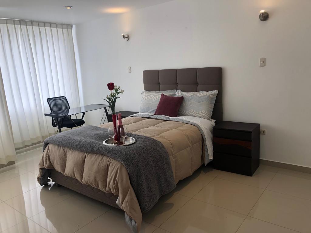 A spacious sleeping area features a comfortable bed layered with neutral bedding and accent pillows. A small table and chair are positioned nearby, with a decorative vase holding a rose. Natural light filters through sheer curtains, enhancing the quiet ambiance of the room.
