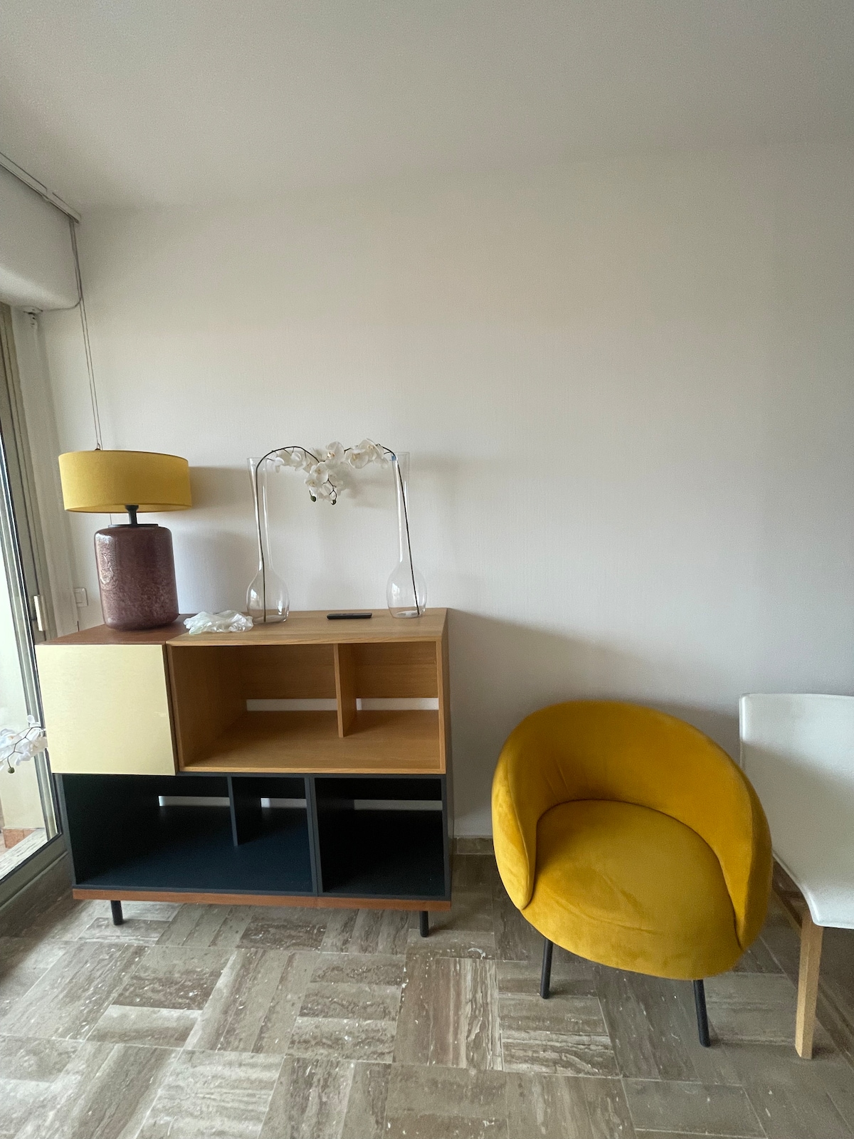 A stylish corner features a decorative lamp placed on a modern cabinet with an open design and a combination of light wood and dark accents. A vibrant yellow chair complements the space, accompanied by a small white table in the background.