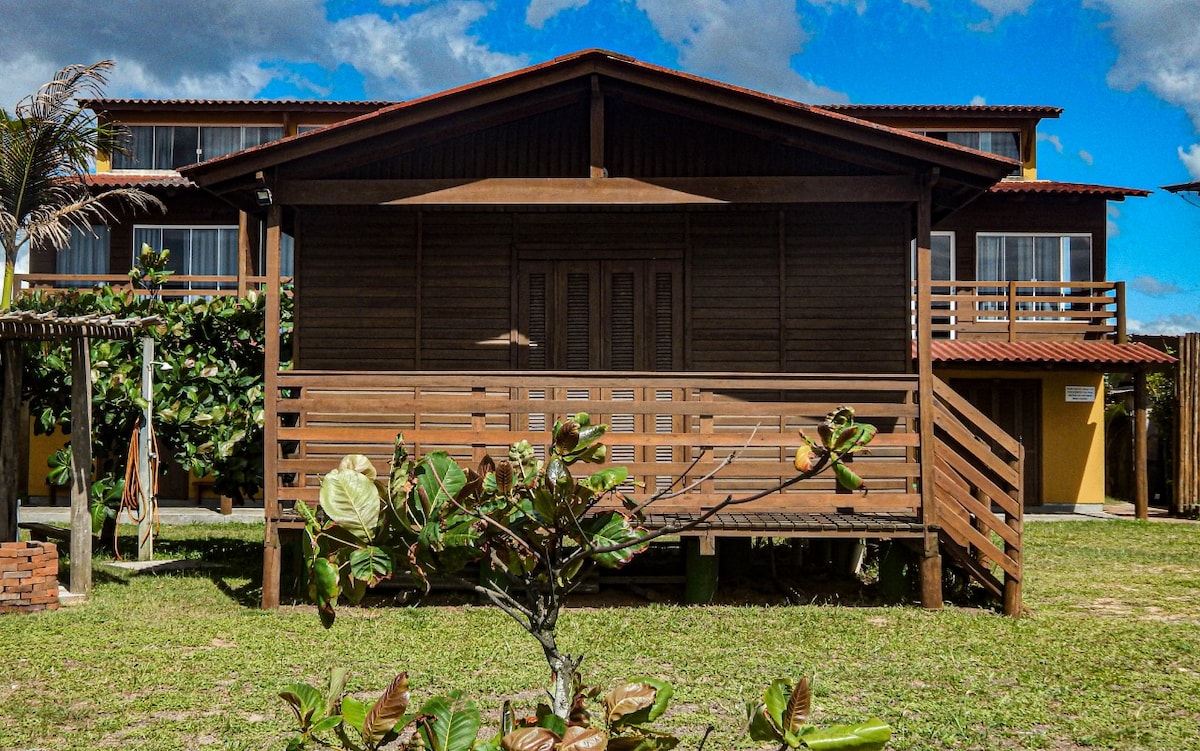 A wooden structure with a sloping roof is set on stilts, featuring a front porch with vertical wood slats for privacy. The building is surrounded by greenery, and the sky is partially cloudy, revealing a sunny atmosphere.