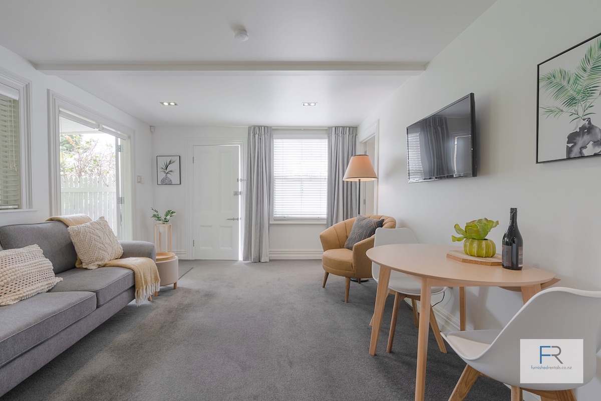 The living area features a comfortable gray sofa and an accent chair. A round dining table is positioned nearby, accompanied by a potted plant and a bottle. Natural light enters through windows, and a mounted television is visible on the wall.