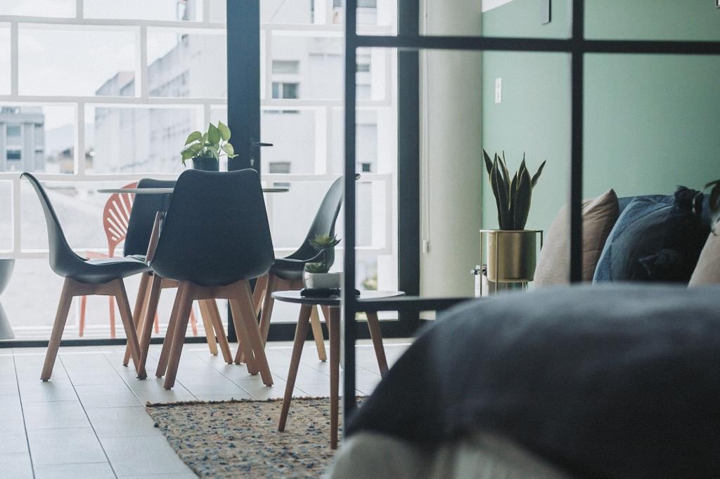 A modern living space is featured, with a small dining area that includes a round table surrounded by four chairs. Houseplants add a touch of greenery, while large windows provide ample natural light, offering a view of the surrounding area.
