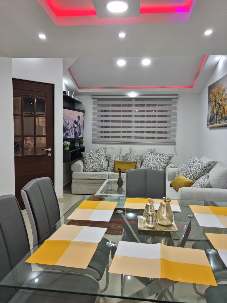 A modern living and dining area is shown, featuring a glass dining table set with neatly arranged place settings. A comfortable light-colored sofa with decorative cushions is positioned against the wall. A television on the shelf adds to the relaxed atmosphere, while soft lighting accents the ceiling.
